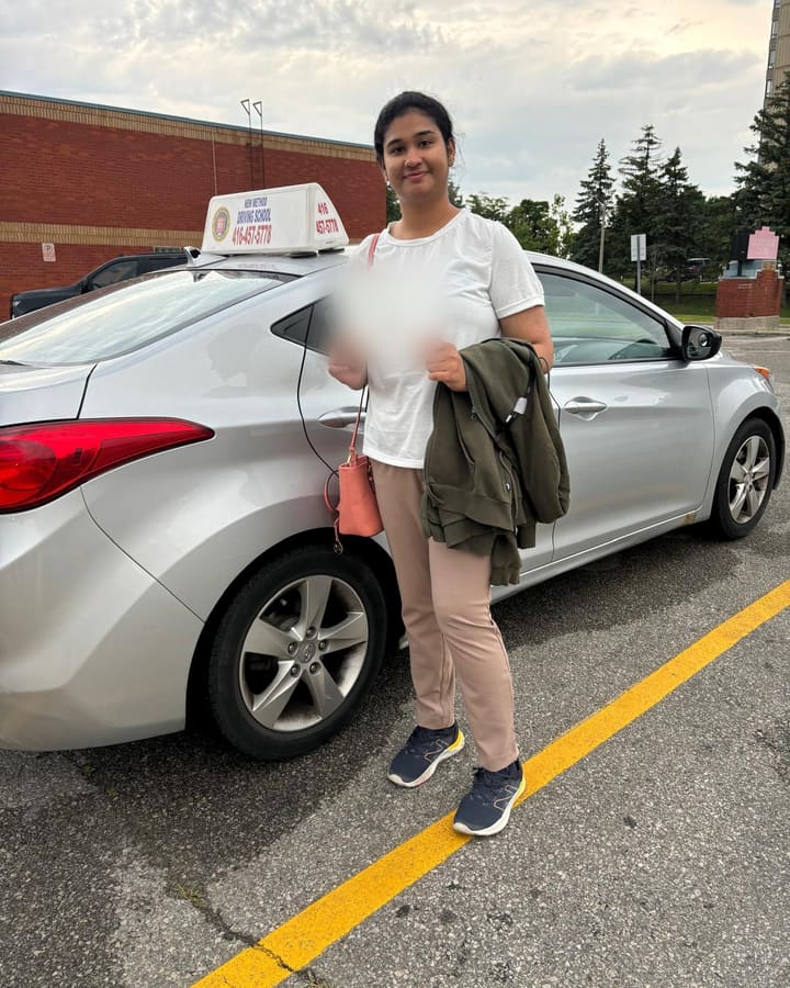 A smiling student of Mrs. Akbar holds her road test paperwork beside a silver lesson car with a driving school sign.